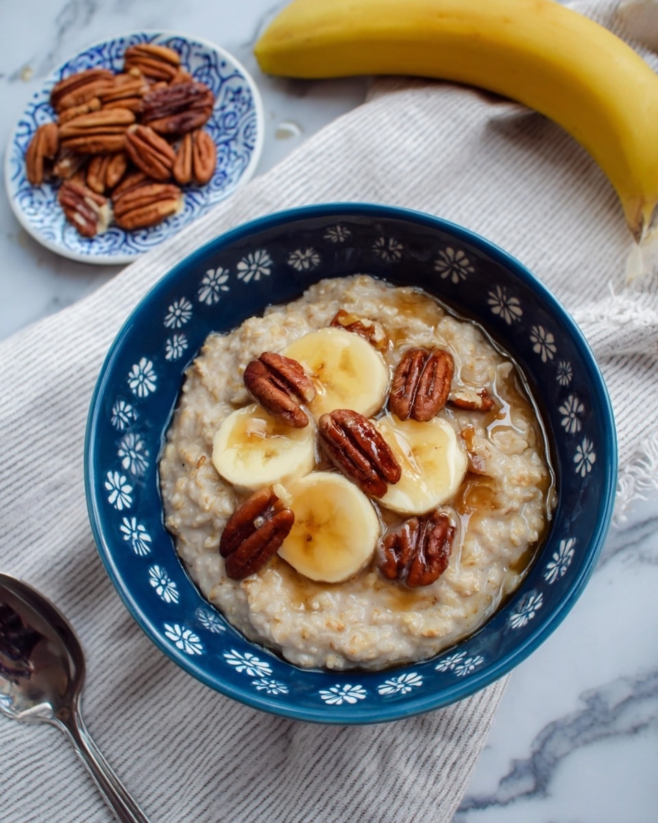 A blue bowl with small white flowers contains a thick oatmeal layer, light beige with a soft, creamy texture, topped with three round yellow banana slices and five dark brown pecans arranged evenly on the banana slices. There is a shiny drizzle of syrup over the oatmeal and toppings. Next to the bowl on a white cloth with gray lines lies a whole yellow banana and a small white plate with blue patterns holding additional pecans. A metal spoon is partially visible at the bottom. The setting is on a white marbled surface. photo taken with an iphone --ar 4:5 --v 7