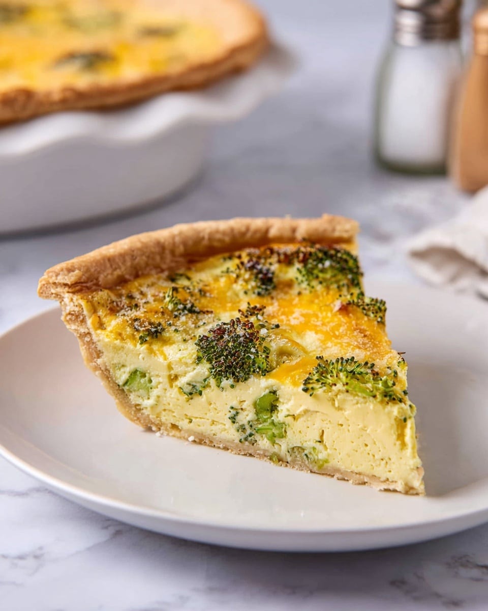 A single slice of broccoli quiche sits centered on a white plate, showing three main layers: a golden brown, flaky crust at the bottom and edges, a light yellow, smooth egg custard filling in the middle with small pieces of broccoli mixed inside, and a top layer featuring bright green broccoli florets and melted yellow cheese with some browned spots. The slice is angled slightly to the right, and behind it, a larger quiche in a white dish is partially visible with a white marbled surface underneath. Salt and pepper shakers stand blurred in the background. photo taken with an iphone --ar 4:5 --v 7