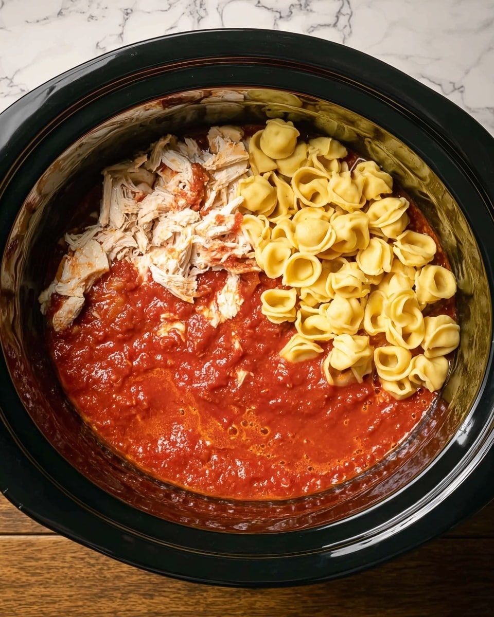 Inside a black slow cooker, there are two main layers: the bottom layer is a thick, red tomato-based sauce with a smooth texture and some bubbles, while the top layer is divided into two parts on one side, there are small pieces of white shredded chicken, and on the other side, a pile of small, yellow cooked tortellini pasta. The slow cooker is placed on a wooden surface but the background is changed to a white marbled texture, photo taken with an iphone --ar 4:5 --v 7