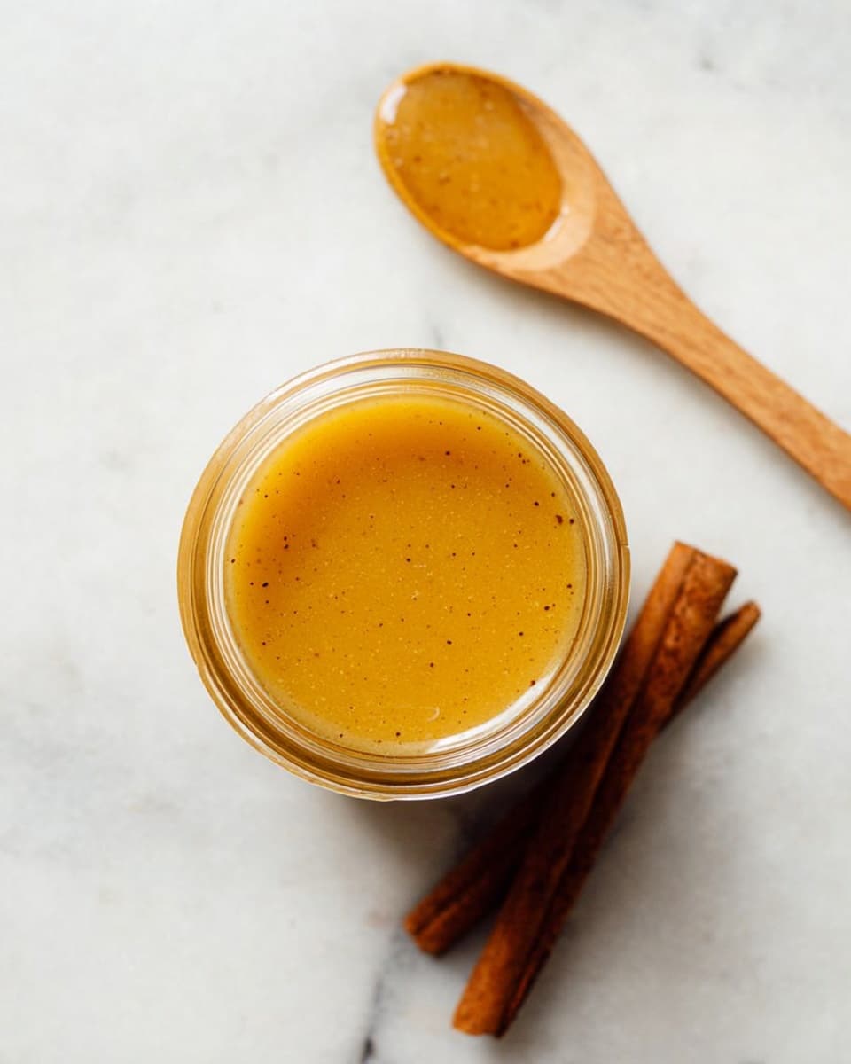 The image shows a small glass jar filled with smooth, golden honey that has tiny dark specks inside it, placed on a white marbled surface. Above the jar, there is a wooden spoon resting diagonally with some honey on it, showing a thick but shiny texture. At the bottom right corner, two cinnamon sticks lie parallel to each other, adding a brown warm tone to the clean background. The photo is shot from above, focusing on the round jar and the natural colors of the honey and cinnamon. photo taken with an iphone --ar 4:5 --v 7