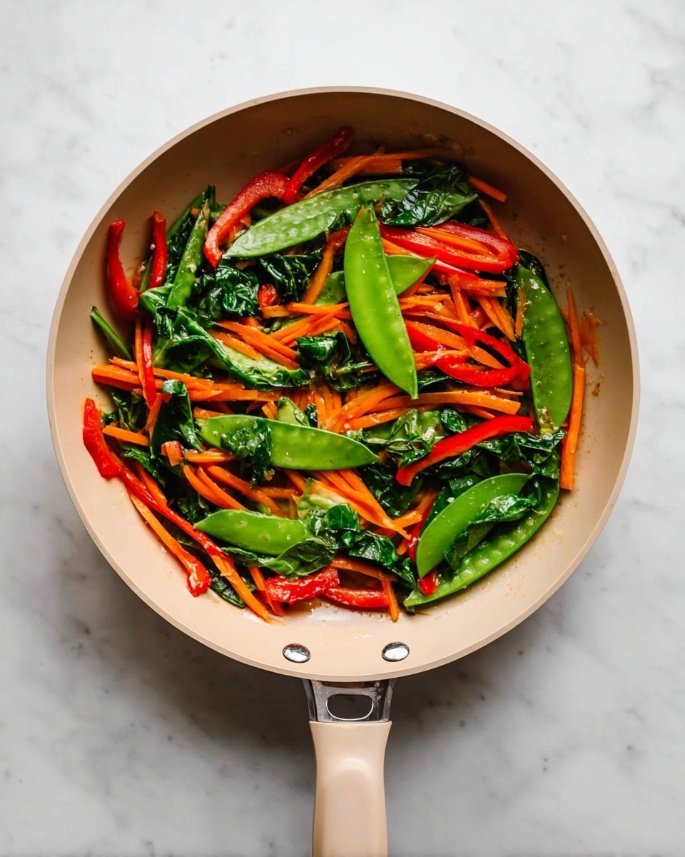 A beige pan placed on a white marbled surface holds a colorful mix of cooked vegetables arranged loosely. The bottom layer is mainly thin orange carrot strips, then bright red bell pepper slices lay scattered across. Large green snow pea pods and dark green leafy spinach pieces spread on top, adding a fresh glossy look. The vegetables have a slight shine from light cooking. The pan’s handle extends downward in the image, with smooth edges and a small round button near the base. photo taken with an iphone --ar 4:5 --v 7