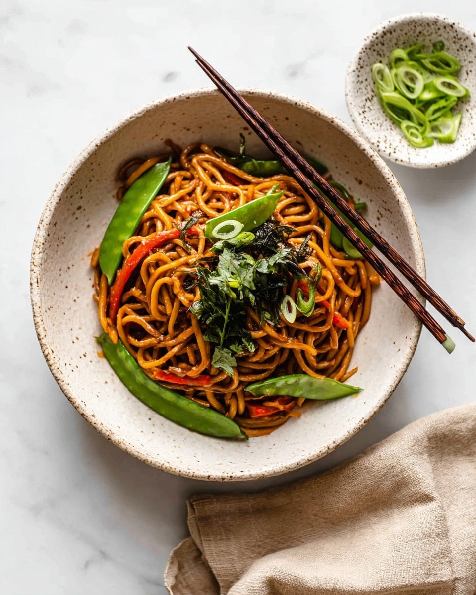 A white ceramic bowl with a speckled, slightly rough texture holds a serving of glossy brown stir-fried noodles mixed with bright green snow peas, thin carrot strips, and garnished with fresh green onion slices and dark green leafy herbs on top. The noodles are thick and slightly twisted, coated evenly with sauce. A pair of dark wooden chopsticks rests on the edge of the bowl. To the upper right, a small white bowl with dark speckles contains more fresh green onion slices. The whole scene is set on a smooth white marbled surface with a soft beige cloth partially visible at the bottom right. Photo taken with an iphone --ar 4:5 --v 7