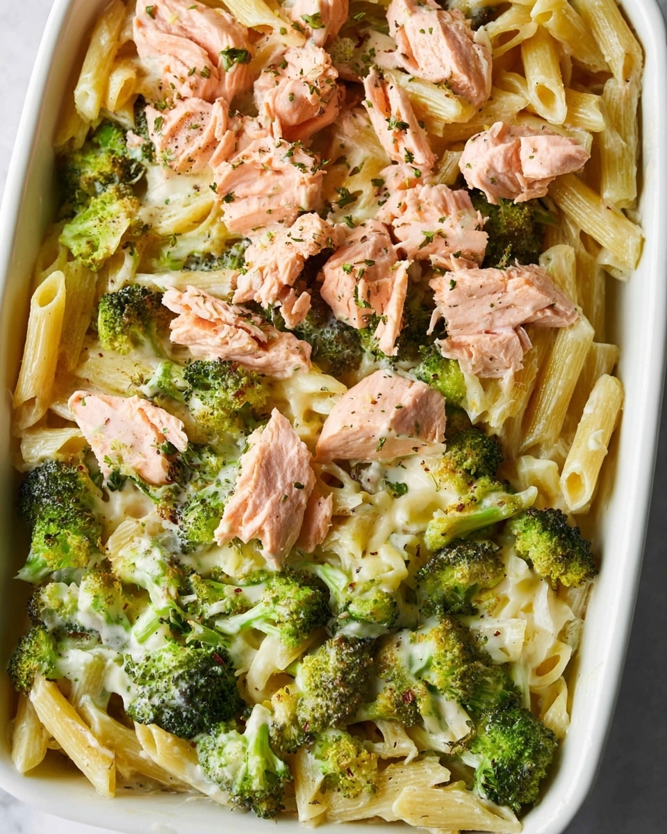 A white rectangular dish holds a layered pasta meal with three main layers: the bottom layer is cooked penne pasta in a pale yellow color, mixed with green broccoli florets that have a slightly roasted texture. On top of this is a creamy light sauce that coats some pasta and broccoli pieces. The top layer features broken pieces of pink cooked salmon, sprinkled with small green herbs and black pepper. The white marbled surface is partially visible around the edges of the dish. photo taken with an iphone --ar 4:5 --v 7