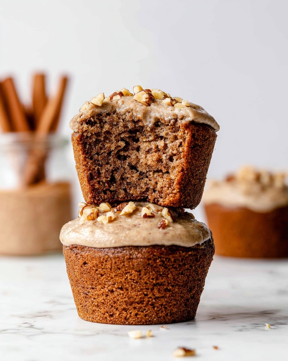The image shows two stacked muffins on a white marbled surface. The bottom muffin is whole, with a brown, slightly textured crust and a light beige frosting on top, sprinkled with small walnut pieces. The top muffin is cut in half, revealing a moist, dense, and crumbly brown inside, while its top half rests on the bottom muffin, showing the same beige frosting with walnut bits. In the blurry background, there is another muffin and a jar with cinnamon sticks inside, all against a clean, pale background. Photo taken with an iphone --ar 4:5 --v 7