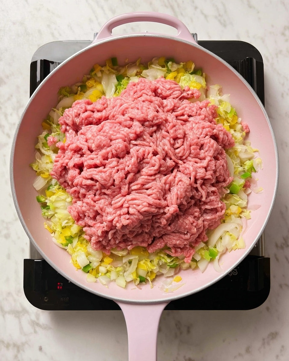 A pink pan filled with a cooking mix is shown, featuring a thick bottom layer of finely chopped yellow, white, and light green vegetables, mostly onions and bell peppers, around the edges. On top of this layer is a large, loose mound of fresh pink ground meat spread across the center, with strands and small chunks visible. The light pink pan has two handles and is placed on a black cooktop, all set on a white marbled surface. Photo taken with an iphone --ar 4:5 --v 7