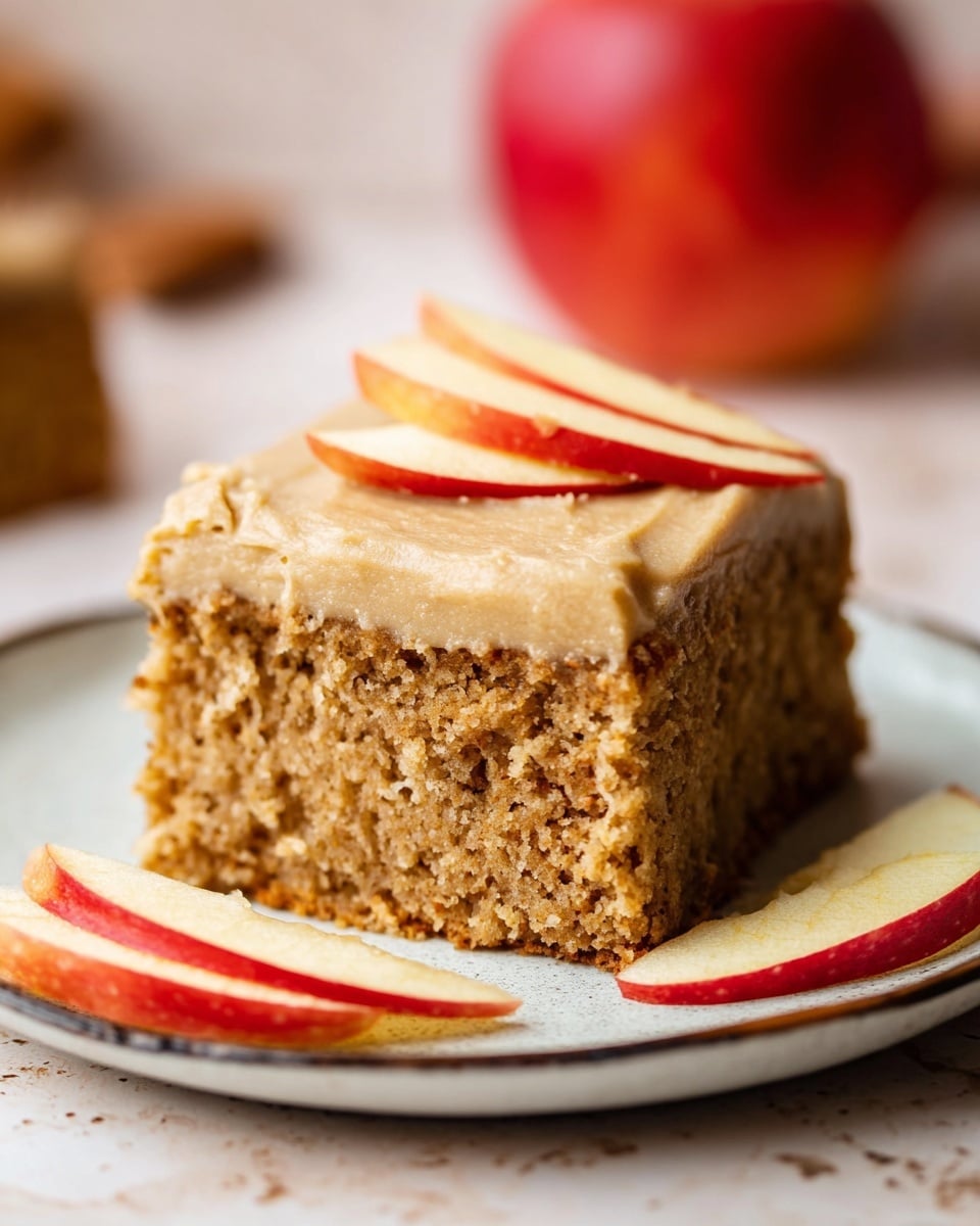 A single square piece of moist, brown apple cake with a rough texture sits on a white plate with a thin dark rim. The cake has one layer covered with a thick light brown frosting on top. Three thin slices of red apple with pale yellow inside rest neatly on the frosting. Next to the cake on the plate are three more apple slices, showing shiny red skin and pale inside. The background has a blurred red apple and a white marbled surface. photo taken with an iphone --ar 4:5 --v 7