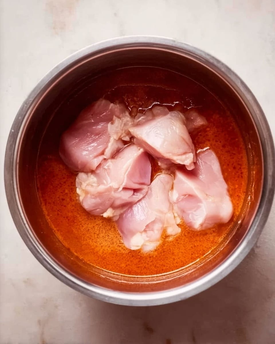 The image shows the inside of a stainless steel pot with an orange-red sauce covering the bottom layer. On top of this sauce, there are several raw pale pink pieces of chicken placed in the center. The sauce has a smooth texture, and the chicken pieces appear fresh and moist with some glossy areas. The pot is shiny and reflects light softly. The background surface around the pot is a white marbled texture. photo taken with an iphone --ar 4:5 --v 7