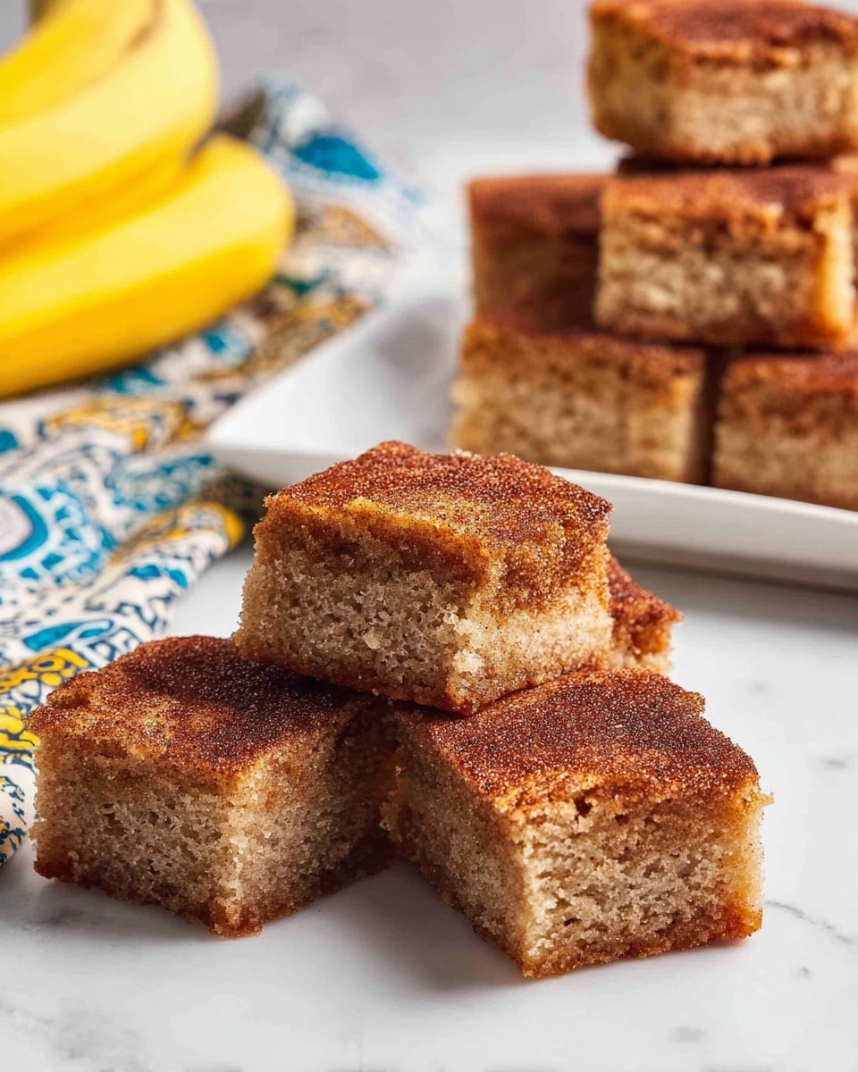 The image shows several square pieces of cinnamon cake with a light brown, moist, and soft texture inside, topped with a thin layer of darker brown cinnamon sugar crust. Three pieces are arranged in a small pile on a white marbled surface, with one piece slightly raised showing the inside crumb. Behind them, more cake squares are stacked on a white rectangular plate, with a yellow banana and a white and blue patterned cloth in the blurred background. Photo taken with an iphone --ar 4:5 --v 7