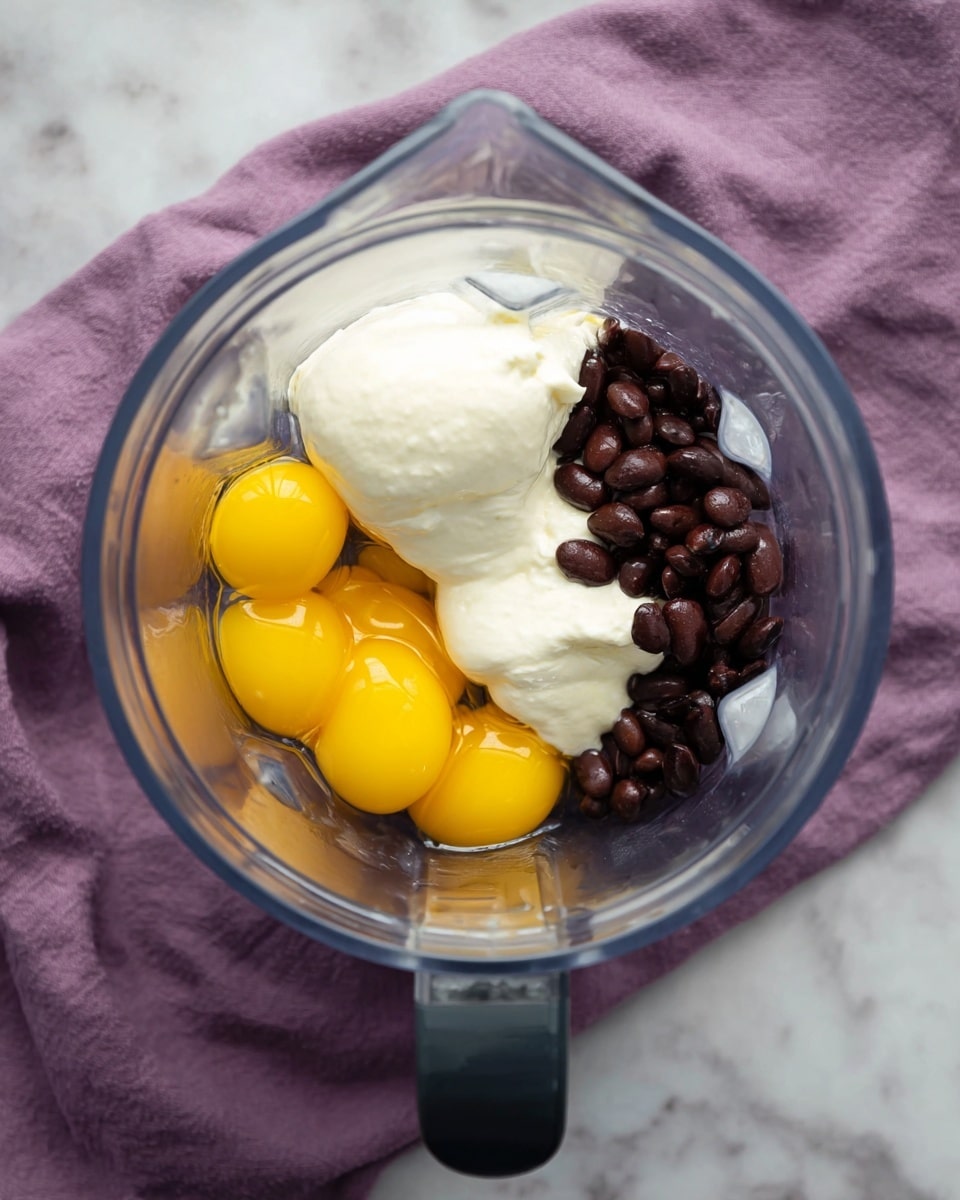 A clear blender jar sits on a white marbled surface with a purple cloth in the background. Inside the blender, there are three bright yellow egg yolks clustered on the left side, smooth white creamy yogurt in the middle, and a pile of dark brown beans on the right side. The ingredients are not mixed, showing clear layers of different textures and colors. The blender jar has a black handle pointing downward. Photo taken with an iphone --ar 4:5 --v 7
