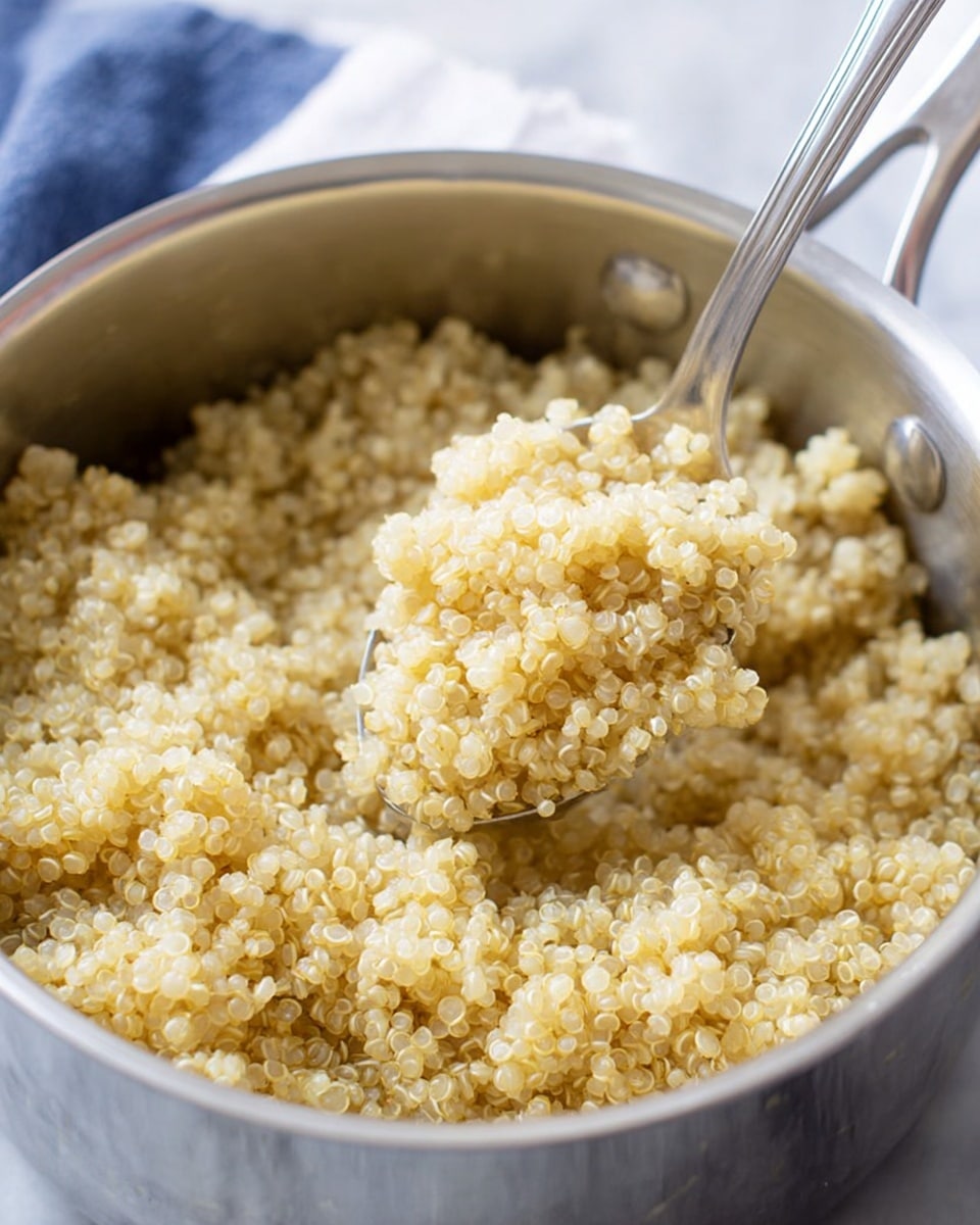 A close-up image of a stainless steel pot filled with light yellow cooked quinoa. The quinoa grains are small, round, and slightly translucent, creating a fluffy texture that fills the pot. A shiny metal spoon is inside the pot, lifting a portion of the quinoa. The background shows a soft white marbled surface with a blurred white and blue cloth. photo taken with an iphone --ar 4:5 --v 7