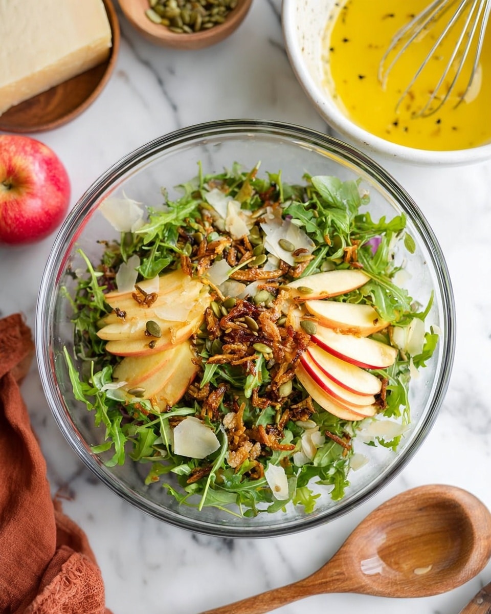 The dish is a clear glass bowl with a fresh salad inside, placed on a white marbled surface. The salad has three main layers: at the bottom, there is a bed of green arugula leaves; the middle layer is made of thinly sliced red and yellow apples arranged in small clusters on top of the greens; scattered on top are crunchy brown fried shallots and green pumpkin seeds. The salad is lightly sprinkled with a dressing that adds a slight shine, and some black pepper bits are visible. Around the bowl, there is a white marbled area with a piece of cheese, a red apple, and a bowl of yellow dressing with a whisk inside. A woman's hand is holding a wooden spoon near the bowl. Photo taken with an iphone --ar 4:5 --v 7