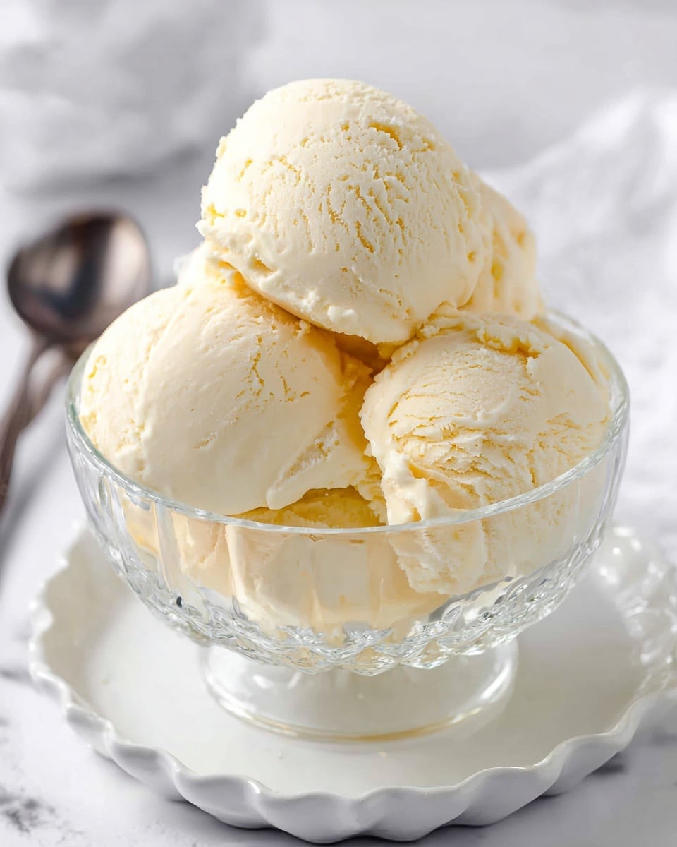 A clear glass bowl filled with five scoops of creamy pale yellow vanilla ice cream, stacked closely together forming a small mound, sitting on a white scalloped dish, all placed on a white marbled surface with a silver spoon in the background slightly out of focus photo taken with an iphone --ar 4:5 --v 7