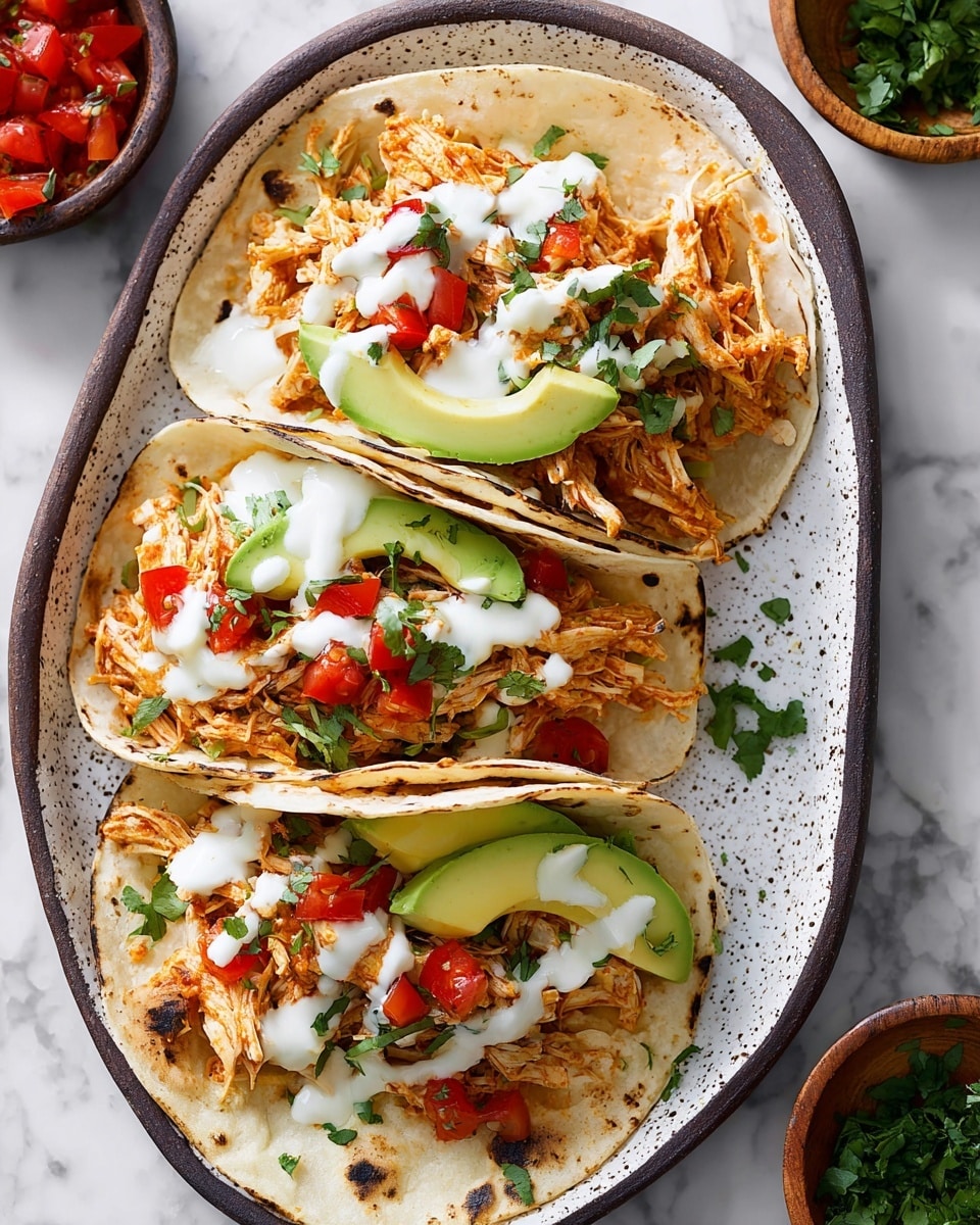 Three soft corn tortillas with light browning and slight charring at edges are layered with shredded orange-hued chicken. Each taco is topped with small red diced tomatoes and fresh green cilantro bits scattered on top. Two tacos have thin, long slices of bright green avocado under the chicken. White creamy sauce is drizzled over each taco in uneven lines. The tacos are neatly placed on a white speckled oval plate with dark brown edges. The background shows a white marbled texture surface, and there are small bowls with chopped herbs and tomatoes partially visible nearby. photo taken with an iphone --ar 4:5 --v 7