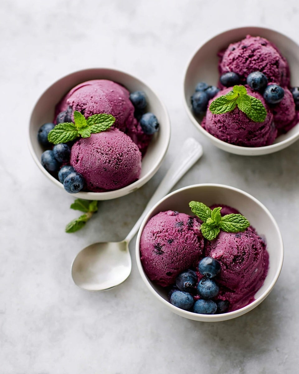 Three white bowls sit on a white marbled surface, each holding three deep purple scoops of ice cream with a smooth but slightly textured surface. Each scoop is paired with a couple of fresh blue blueberries and a few green mint leaves, providing color contrast. A white spoon rests inside the closest bowl on the left side. The bowls are arranged in a triangular formation with soft natural light highlighting the colors. Photo taken with an iphone --ar 4:5 --v 7