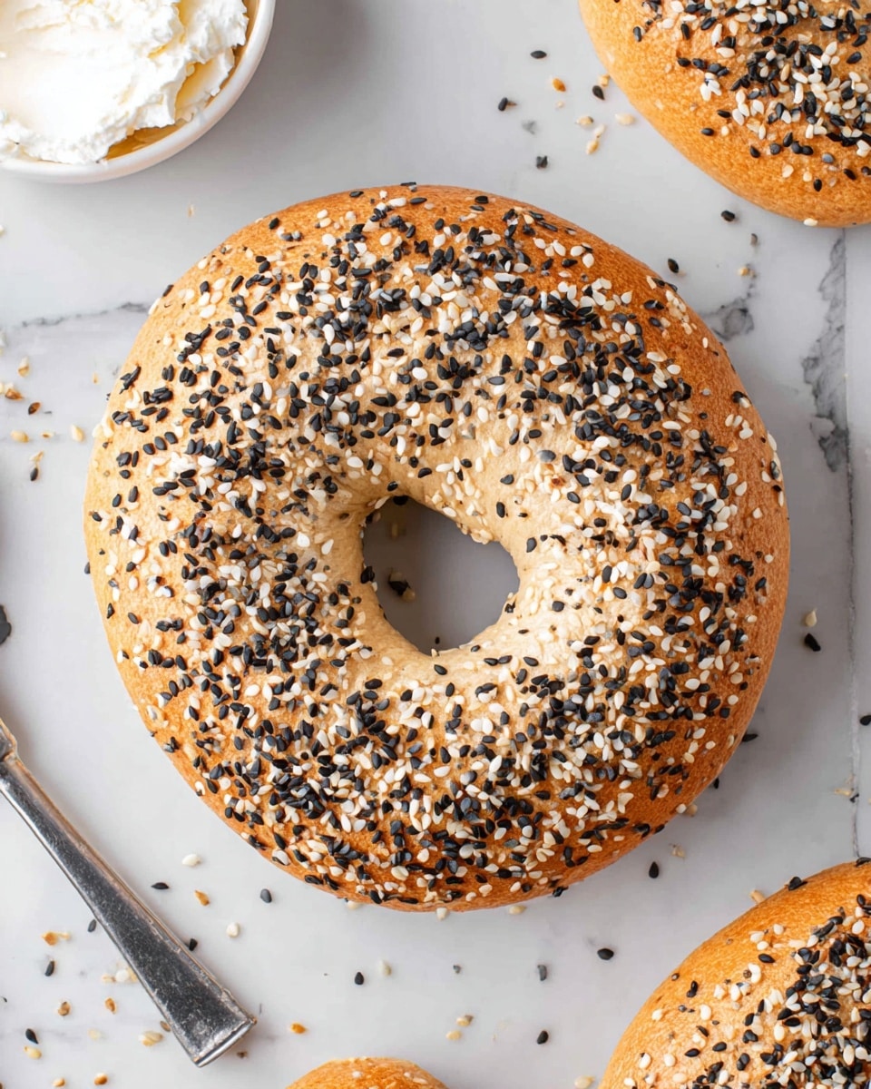 The image shows a golden brown bagel covered with a generous layer of white and black sesame seeds all over the top. The bagel’s surface is smooth and slightly shiny, with a firm texture visible. It is placed on a white marbled surface with scattered sesame seeds around it. The bagel has a round shape with a large hole in the center. In the corner, part of a white bowl with a creamy white spread and a silver knife is visible. photo taken with an iphone --ar 4:5 --v 7