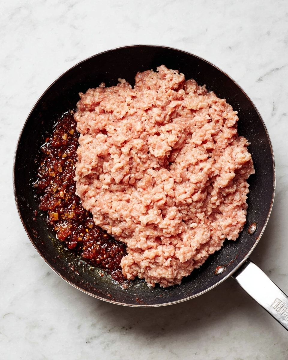 A shallow black pan with a white handle sits on a white marbled surface, containing two layers: the bottom layer is a dark brown finely chopped mixture with a shiny, oily texture, mostly covering the lower left section of the pan; the top layer is a thick, pale pinkish raw ground meat spread with a rough texture, mostly covering the upper and right parts of the pan, with some meat overlapping the brown mixture along the bottom edge. photo taken with an iphone --ar 4:5 --v 7
