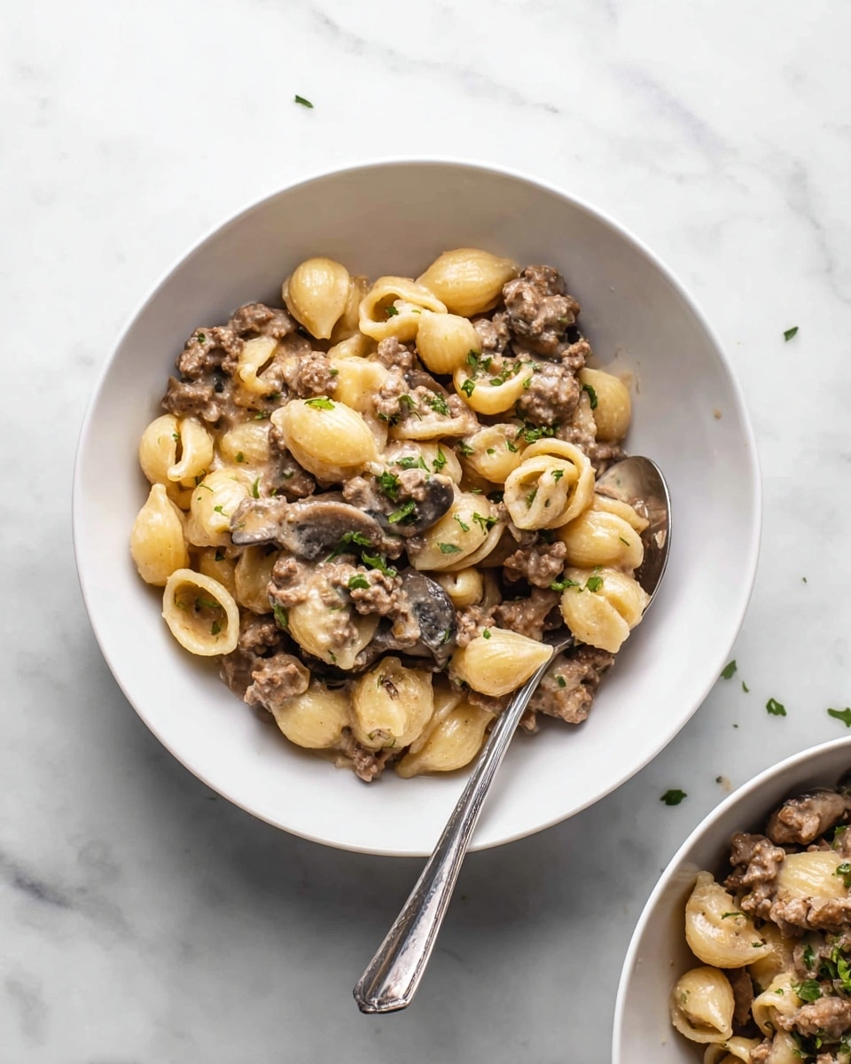 Two white bowls are placed on a white marbled surface, each filled with small shell pasta mixed with ground meat and mushroom slices in a creamy sauce. The pasta is yellowish and shaped like little shells, while the meat is brown and crumbly, and the mushrooms are sliced and darker. Small green herb pieces are sprinkled on top for color. One bowl is near the center with a silver spoon resting inside, and the other bowl is partially visible in the upper right corner. The photo taken with an iphone --ar 4:5 --v 7