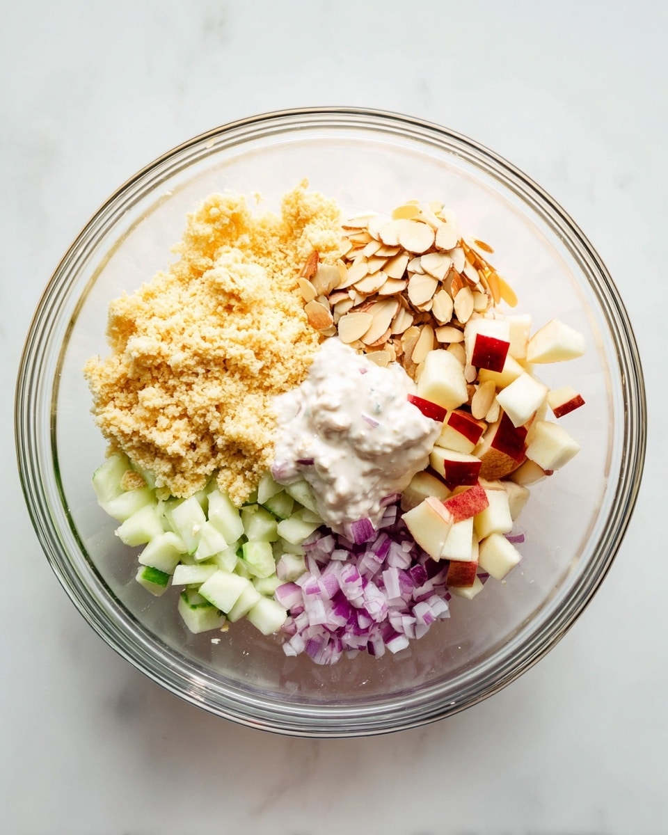 A clear glass bowl contains six main layers arranged separately inside it. On the left side, there is a large portion of light yellow crumbly texture. Next to it on the top right side, there are thin slices of light brown almonds. Below the almonds, there are small pieces of red and white apple cubes. Below the apples, there are small pale green cucumber cubes. Next to the cucumber, on the bottom left, there are small purple-red onion cubes. In the middle of the bowl, resting over the apples and close to the crumbly layer, there is a white creamy sauce. The bowl is placed on a white marbled surface photo taken with an iphone --ar 4:5 --v 7