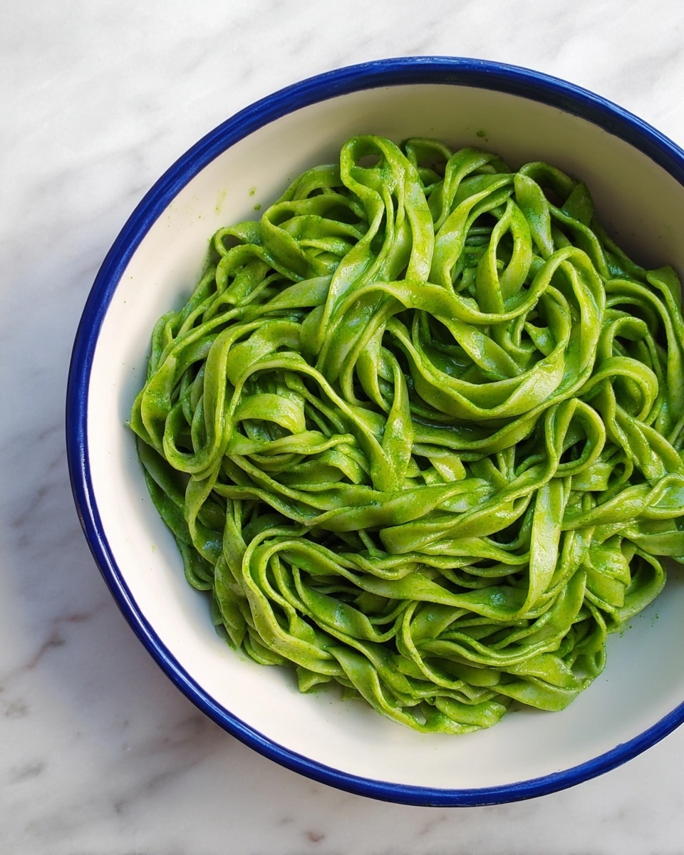 A white bowl with a blue rim holds one layer of green pasta noodles that are twisted and slightly shiny. The pasta has a rough texture and a thick width, filling the bowl without any extra toppings. The bowl sits on a white marbled surface. Photo taken with an iphone --ar 4:5 --v 7