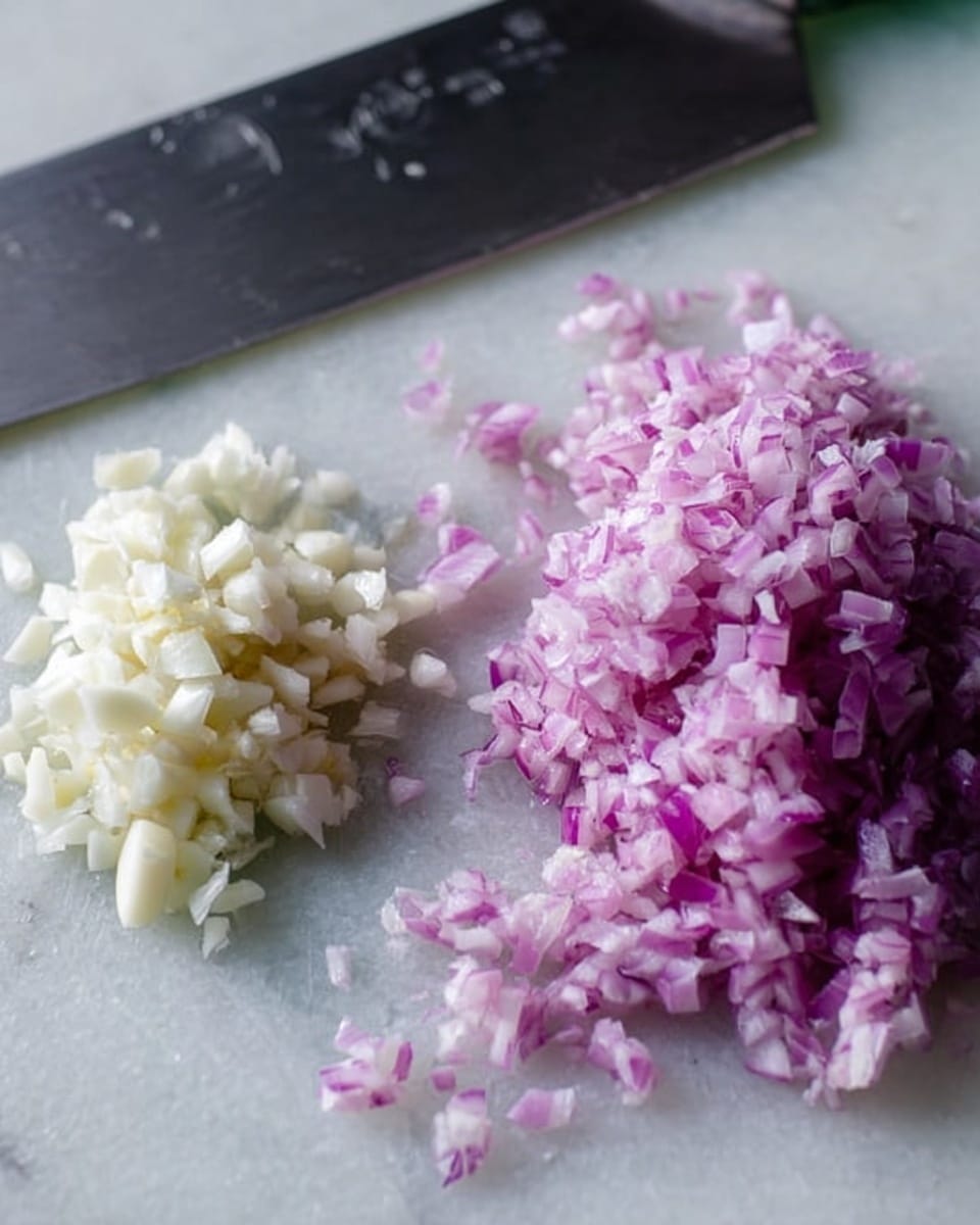 The image shows two piles of finely chopped ingredients on a white marbled surface. On the left, there is a small pile of chopped garlic, which is off-white with a soft texture. On the right, there is a larger pile of chopped red onion, showing a light purple color with a slightly translucent texture. Behind the piles, there is a large stainless steel knife resting flat on the surface, with the blade close to the chopped onion. photo taken with an iphone --ar 4:5 --v 7