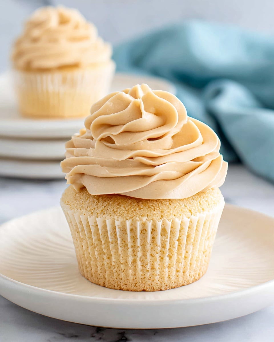 A light yellow cupcake sits centered on a white plate with subtle ridges. The cupcake has a soft texture with a slightly golden base. On top, there is a large swirl of smooth, creamy frosting in a pale tan color, piped in multiple rounded layers forming a flower-like shape. In the background, another cupcake on a white plate is softly blurred, with a light blue cloth adding a gentle color contrast behind it, all set on a white marbled surface. photo taken with an iphone --ar 4:5 --v 7