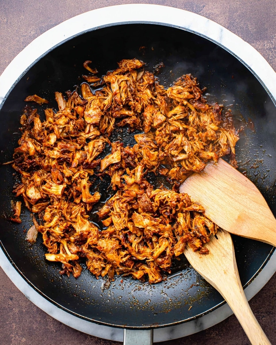 A black pan is filled with shredded, cooked jackfruit pieces that have a mix of dark brown and orange colors, showing a slightly crispy and saucy texture. The jackfruit sits unevenly in the pan, with some pieces clumped together and others spread out. A light-colored wooden spatula is placed on the right side of the pan, partially covered with the saucy jackfruit. The pan rests on a round white marbled surface, and faint steam rises gently from the food. photo taken with an iphone --ar 4:5 --v 7
