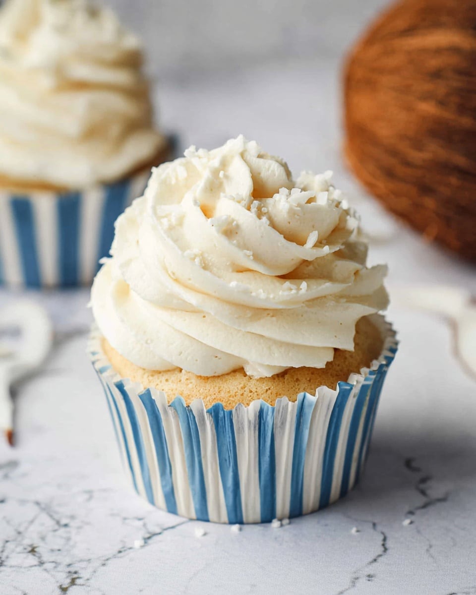 A single cupcake in a blue and white striped paper cup sits on a white marbled surface. The cupcake has a light golden base topped with a thick, creamy white frosting that swirls upwards in multiple layers, creating a soft, textured peak. The frosting looks smooth and slightly fluffy, with small white sprinkles on top adding extra detail. In the background, another cupcake with similar frosting is partially visible, along with a brown coconut resting out of focus. Photo taken with an iphone --ar 4:5 --v 7