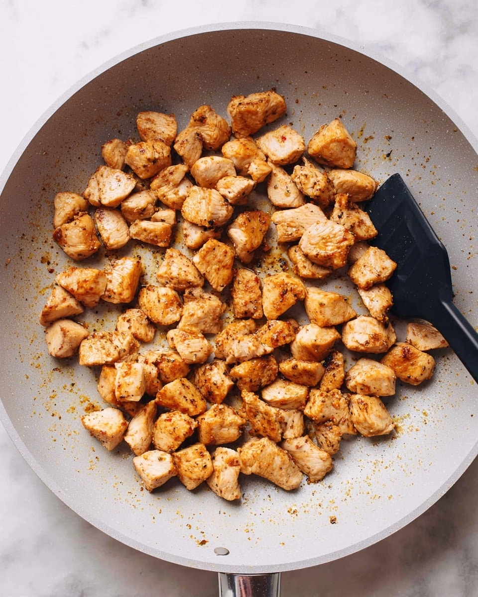 Small golden-brown cooked chicken pieces are spread across a light gray frying pan. The chicken pieces have a slightly crispy outside with a soft texture inside, showing hints of seasoning. A black cooking tool is resting on the right side of the pan. The pan is placed on a surface with a white marbled texture. Photo taken with an iphone --ar 4:5 --v 7