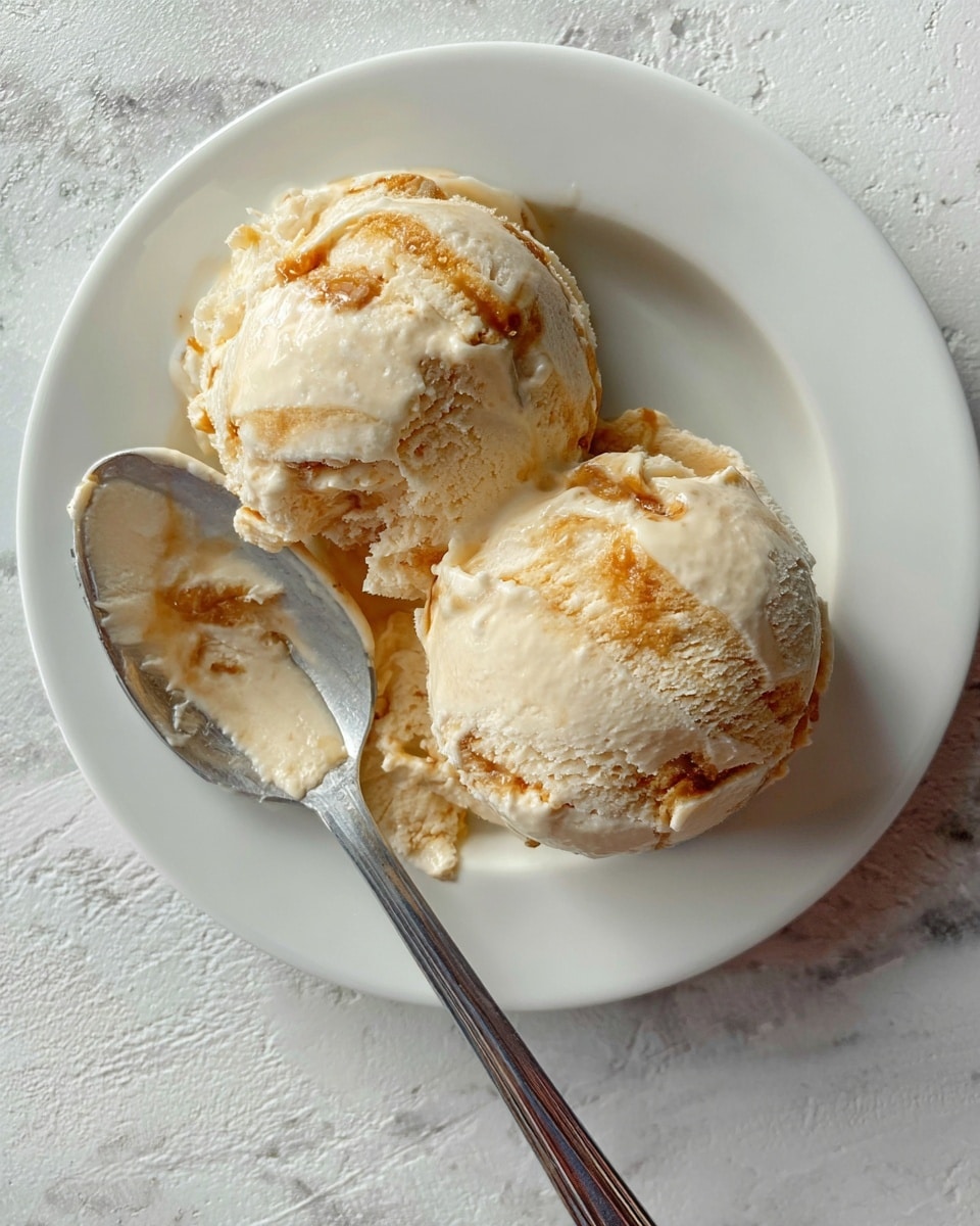Two large scoops of creamy light beige ice cream with golden caramel swirls are placed on a white plate. The ice cream has a soft texture with visible caramel streaks on top and mixed inside. A shiny metal spoon with some ice cream on it rests on the plate next to the scoops. The plate sits on a white marbled surface. Photo taken with an iphone --ar 4:5 --v 7