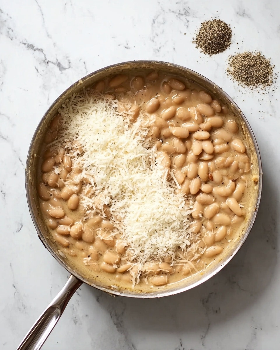 A round stainless steel pan filled with light beige cooked beans in thick sauce, topped with a pile of white grated cheese on one side and sprinkled with ground black pepper nearby, all placed on a white marbled surface. photo taken with an iphone --ar 4:5 --v 7