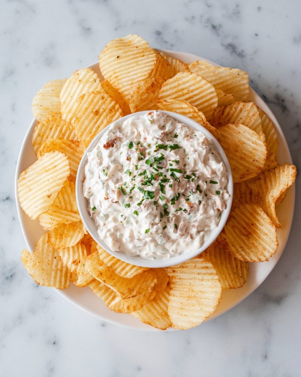 A white plate filled with a ring of golden ridged potato chips surrounding a white bowl placed in the center, which contains a thick creamy dip mixed with small light brown bits and topped with a sprinkle of chopped green herbs. The plate sits on a white marbled surface. photo taken with an iphone --ar 4:5 --v 7