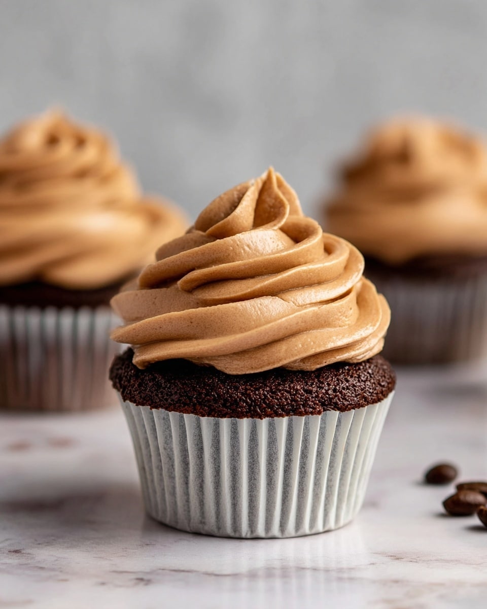 A close-up of a chocolate cupcake with one main layer of dark, moist cake at the bottom, covered by a thick swirl of light brown, smooth frosting on top, creating soft waves and folds. The cupcake is wrapped in a white paper liner with vertical ridges. In the background, two more cupcakes with the same colors and frosting style are slightly out of focus, placed on a white marbled surface with a few coffee beans scattered nearby. The lighting is soft, highlighting the texture of the frosting and cake. Photo taken with an iphone --ar 4:5 --v 7