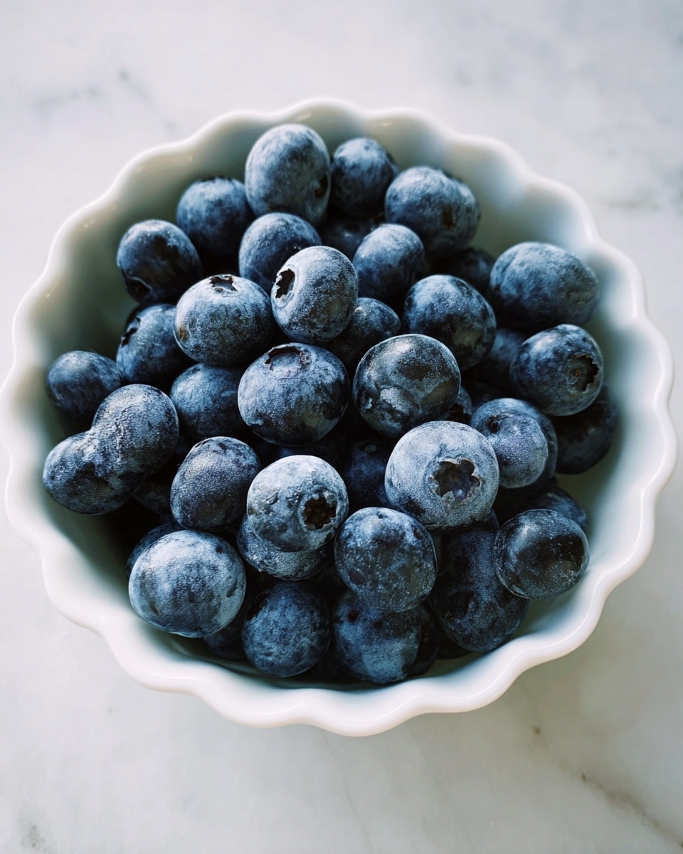 A white scalloped bowl filled with many fresh blueberries, deep blue with a slight frost-like texture on their surface, tightly packed and slightly heaped above the rim, sitting on a white marbled surface with soft natural light casting gentle shadows. photo taken with an iphone --ar 4:5 --v 7