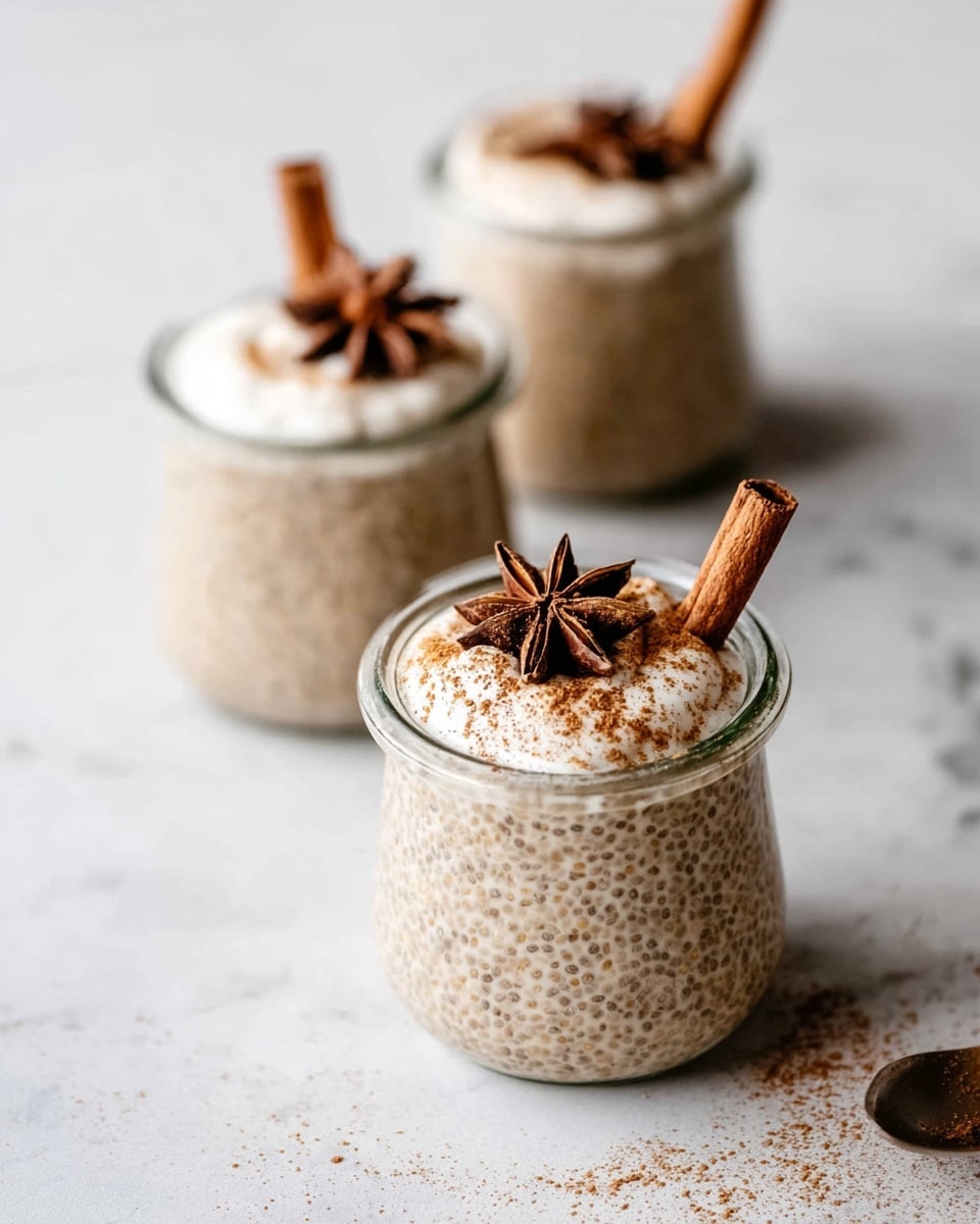 The image shows three small glass jars of chia seed pudding placed on a white marbled surface. Each jar is filled with light beige chia pudding with visible tiny seeds throughout. On top of each pudding is a dollop of creamy white topping, sprinkled with fine brown powder, likely cinnamon. Each jar is garnished with a single cinnamon stick and a star anise, with one jar in the middle being the most in focus and closer to the camera. The overall look is clean and natural with warm earthy tones. Photo taken with an iphone --ar 4:5 --v 7