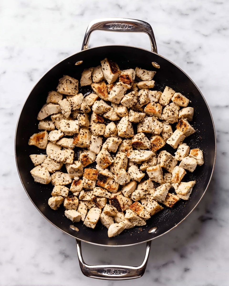 A black pan with two silver handles on each side holds many small, light beige pieces of cooked chicken that have browned edges and are sprinkled with black pepper. The chicken pieces are evenly spread inside the pan, showing a slightly crispy texture with some areas lightly golden. The pan sits on a white marbled surface, which adds a clean background to the simple, cooked chicken pieces. photo taken with an iphone --ar 4:5 --v 7