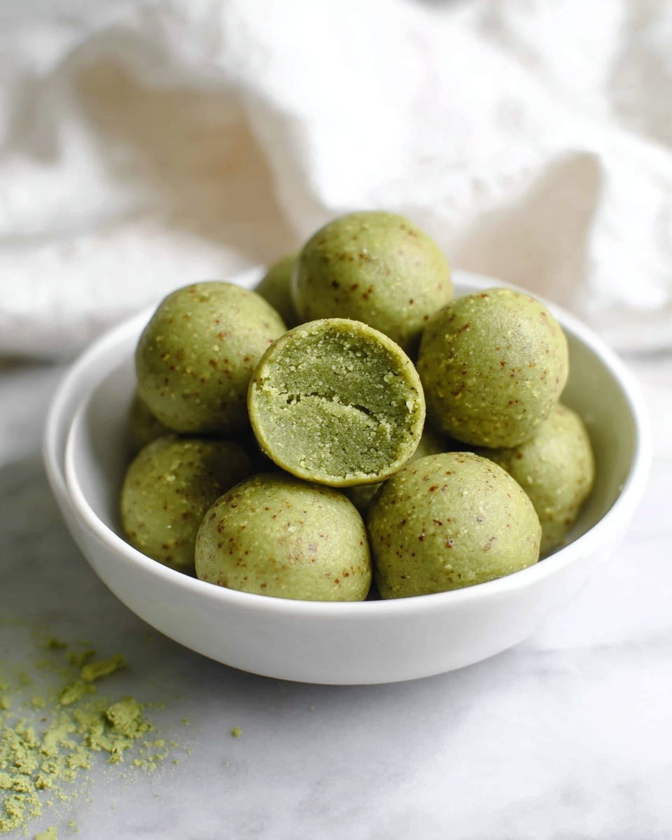 The image shows a white bowl filled with round green balls that have a smooth outer surface with small specks of darker and lighter green. One ball is bitten in half, revealing a layered green interior that looks soft and dense with tiny bits inside. The bowl is placed on a white marbled surface with a soft white cloth in the background. photo taken with an iphone --ar 4:5 --v 7