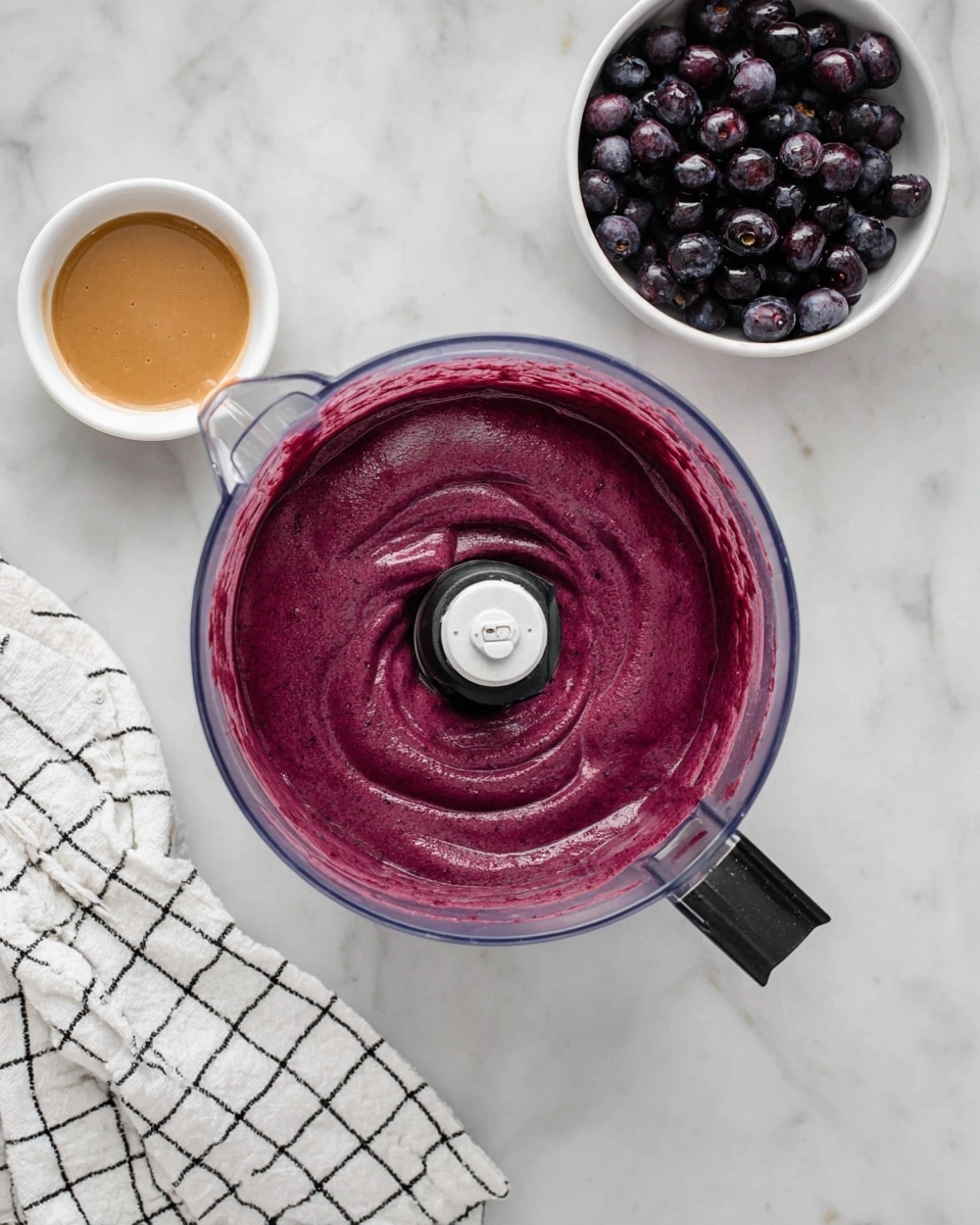 A clear food processor bowl filled with smooth, thick purple berry puree with some light swirls and the blade visible in the center; above it on a white marbled surface are two small white bowls, one filled with dark purple whole berries and the other with a light brown liquid; a white kitchen towel with a black grid pattern lies partially folded in the lower left corner; photo taken with an iphone --ar 4:5 --v 7