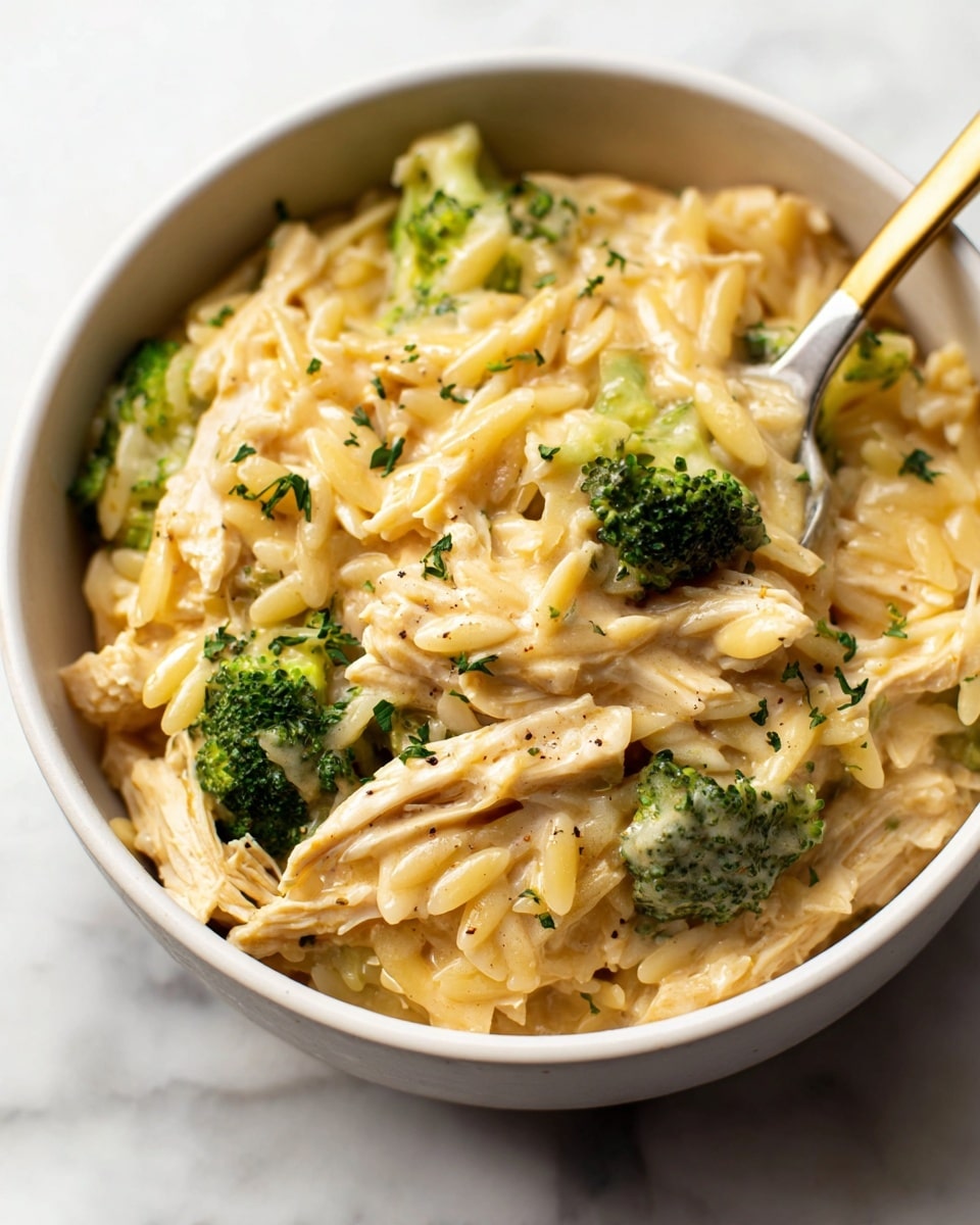 A close-up view of a creamy dish in a white bowl featuring three main layers mixed together: shredded light yellow chicken pieces, small green broccoli florets, and small, oval-shaped pale yellow orzo pasta. The dish is covered with a smooth, rich, beige cheese sauce that coats all ingredients evenly, sprinkled with small bits of green herbs and black pepper for texture. A spoon with a golden head and white handle is partially dipped into the bowl, scooping up some of the creamy mix. The bowl sits on a white marbled surface. photo taken with an iphone --ar 4:5 --v 7