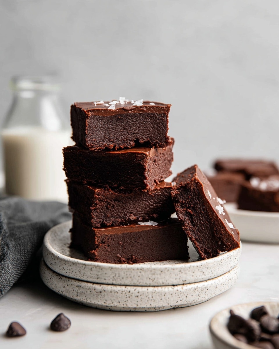 A stack of five thick, dark brown chocolate brownies sits on a white speckled plate, which is placed on another matching plate. The brownies have a dense, slightly crumbly texture and a smooth, glossy chocolate layer on top. The top brownie has small white flakes sprinkled on it. One brownie is leaning against the stack, showing the rich, moist inside. There are a few chocolate chips scattered on the white marbled surface around the plates, with a glass bottle of milk blurred in the background. photo taken with an iphone --ar 4:5 --v 7