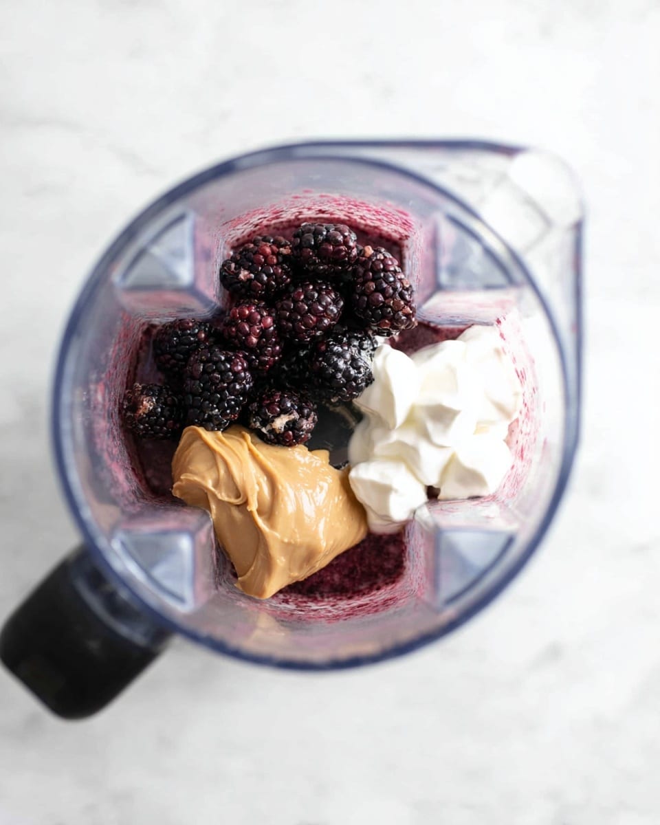 Inside a clear blender jug placed on a white marbled surface, there are four main layers of ingredients. The bottom layer shows a little bit of purple-red liquid. Above that, on the left side, a cluster of dark purple and black small berries, including blackberries and blueberries, forms a textured pile. On the right side, there are two white dollops of thick cream or yogurt. In front of the white dollops, a smooth, light brown peanut butter scoop rests prominently. The blender’s blades and jug edges frame these ingredients from above. Photo taken with an iphone --ar 4:5 --v 7