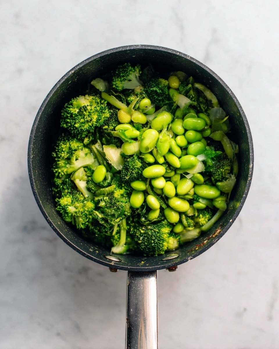 A black cooking pot filled with two main layers of bright green vegetables: tender broccoli florets and smooth edamame beans, all resting evenly inside the pot with visible water drops giving a fresh, wet look. The pot has a long metal handle extending outward and it sits on a white marbled surface. photo taken with an iphone --ar 4:5 --v 7