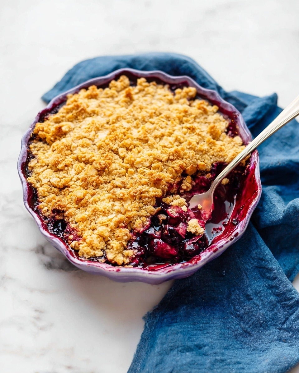 The image shows a round purple ceramic pie dish filled with a fruit crumble dessert. The dessert has two layers: the bottom layer is a deep purple fruit filling, likely berries, which looks juicy and thick, and the top layer is a golden-brown crumbly oat and flour crust, unevenly spread and crunchy in texture. A silver spoon is inside the dish, resting in the empty space where a portion has been scooped out, showing the rich fruit layer underneath. The dish sits on a blue cloth on a white marbled surface. photo taken with an iphone --ar 4:5 --v 7
