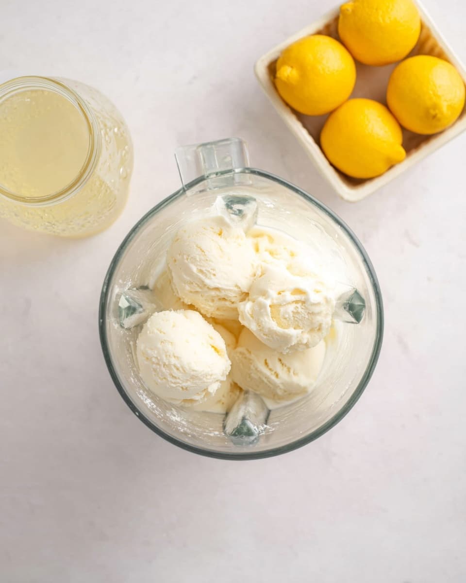In the clear glass blender jar at the center, there are four large scoops of creamy white ice cream with a soft and smooth texture. To the upper left of the blender, a glass jar filled with a pale yellow liquid is placed, showing some condensation on its sides. In the upper right corner, a white container holds four whole bright yellow lemons with a slightly shiny skin. The items all rest on a white marbled surface that gives a clean and bright look to the scene. photo taken with an iphone --ar 4:5 --v 7