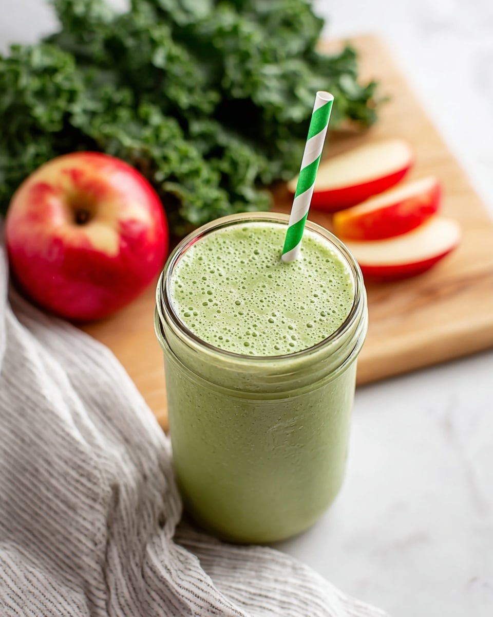 A glass jar filled with a smooth light green smoothie, showing tiny bubbles and a creamy texture on top, with a green and white striped paper straw standing upright in the center. Behind the jar, there are fresh dark green kale leaves and three red apple wedges arranged on a light wooden board. A white cloth with thin black stripes is draped near the bottom left corner of the frame, all set on a white marbled surface. photo taken with an iphone --ar 4:5 --v 7