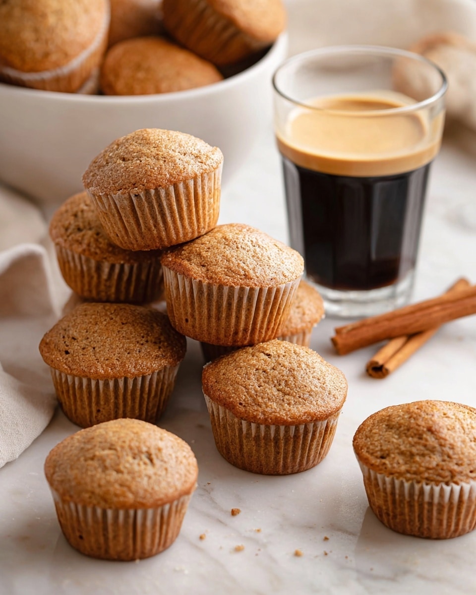 The image shows a group of light brown muffins with a soft, slightly bumpy texture, some stacked on top of each other and others spread loosely on a white marbled surface. Behind the muffins is a clear glass of dark espresso with a light brown creamy layer on top. To the side there are two cinnamon sticks, adding a warm touch, and in the background, extra muffins are placed in a white bowl. The overall scene is bright with natural light, highlighting the warm tones of the muffins and coffee. photo taken with an iphone --ar 4:5 --v 7