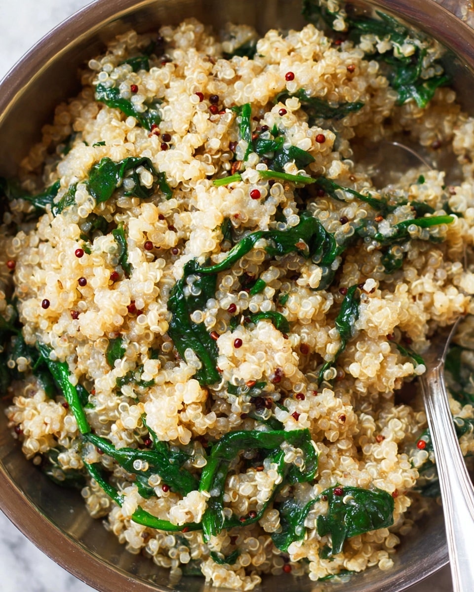 The image shows a close-up of a creamy quinoa dish mixed with cooked spinach leaves. The quinoa is light beige with small fluffy texture and scattered red quinoa seeds, while the spinach is dark green with soft, slightly wilted leaves. The mixture is in a metal bowl with a spoon partially visible on the right side, resting within the grain and spinach blend. The background is a white marbled surface, enhancing the natural colors of the dish. photo taken with an iphone --ar 4:5 --v 7