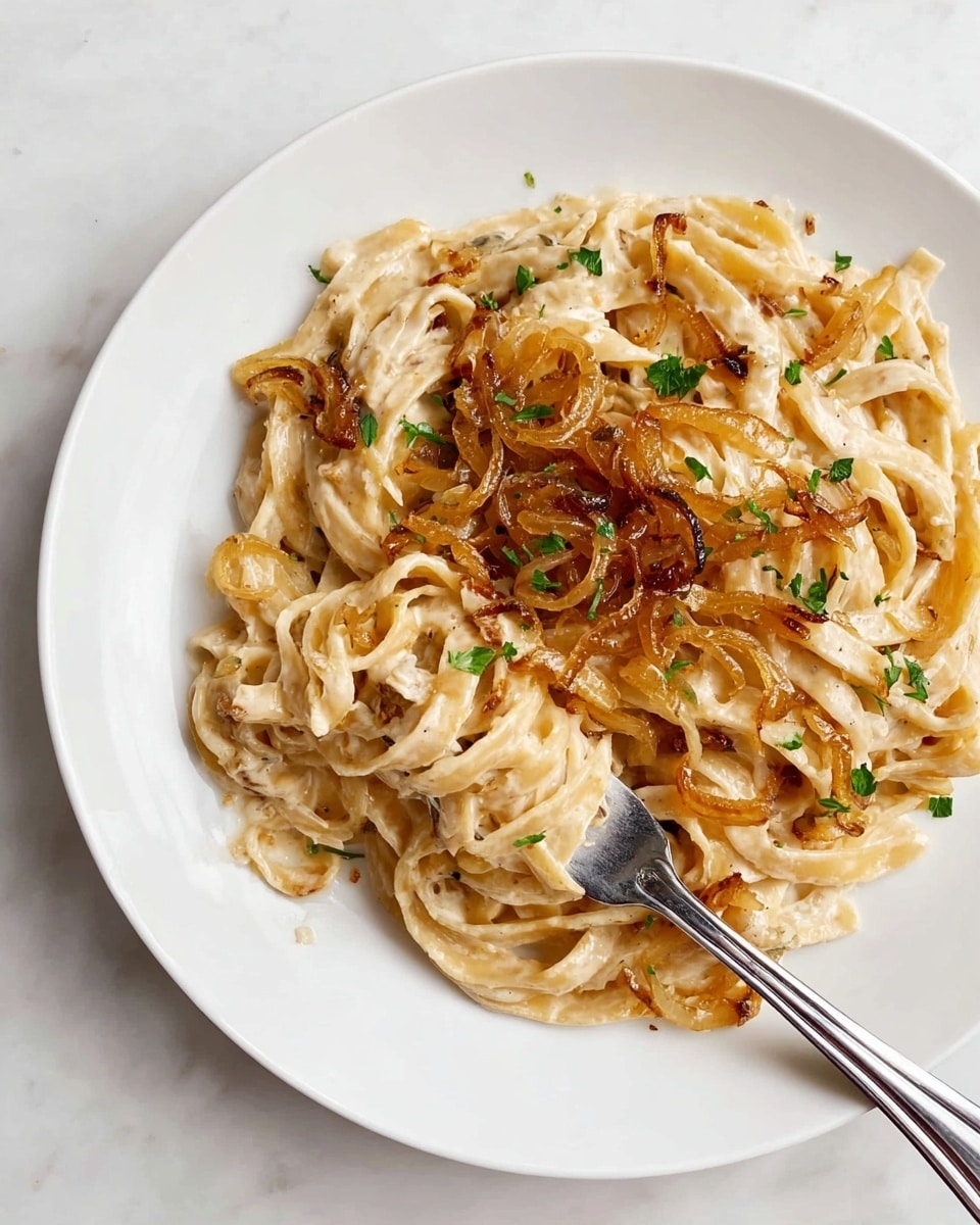 A plate of creamy pasta with three layers: the bottom layer is a white sauce coating long, flat fettuccine noodles in a pale beige color, the middle layer consists of golden-brown caramelized onions spread unevenly on top, and the top layer is small green herb leaves scattered for garnish. A silver fork is placed in the middle right, twirling a few noodles around its tines. The dish is served on a white round plate with a wide rim, set on a white marbled surface. Photo taken with an iphone --ar 4:5 --v 7