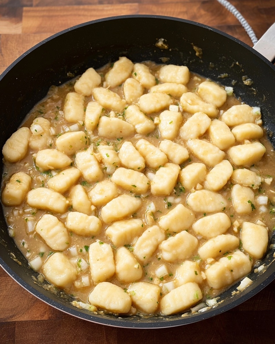 A black pan is filled with small, soft, pale yellow gnocchi shapes floating on a light brown sauce that has small bits of green herbs and white onions visible around the edges. The gnocchi pieces are evenly spread and appear soft and pillowy, covering most of the sauce beneath. The pan sits on a wooden surface with a small part of a white cord visible in the top right corner. The background is a white marbled texture photo taken with an iphone --ar 4:5 --v 7