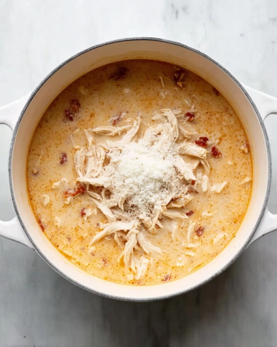 A white pot filled with creamy, light orange soup with visible small pieces of red sun-dried tomatoes and shredded white chicken on top; there is a pile of white powder, likely flour, sprinkled on the chicken in the center, and the pot is placed on a white marbled surface. photo taken with an iphone --ar 4:5 --v 7