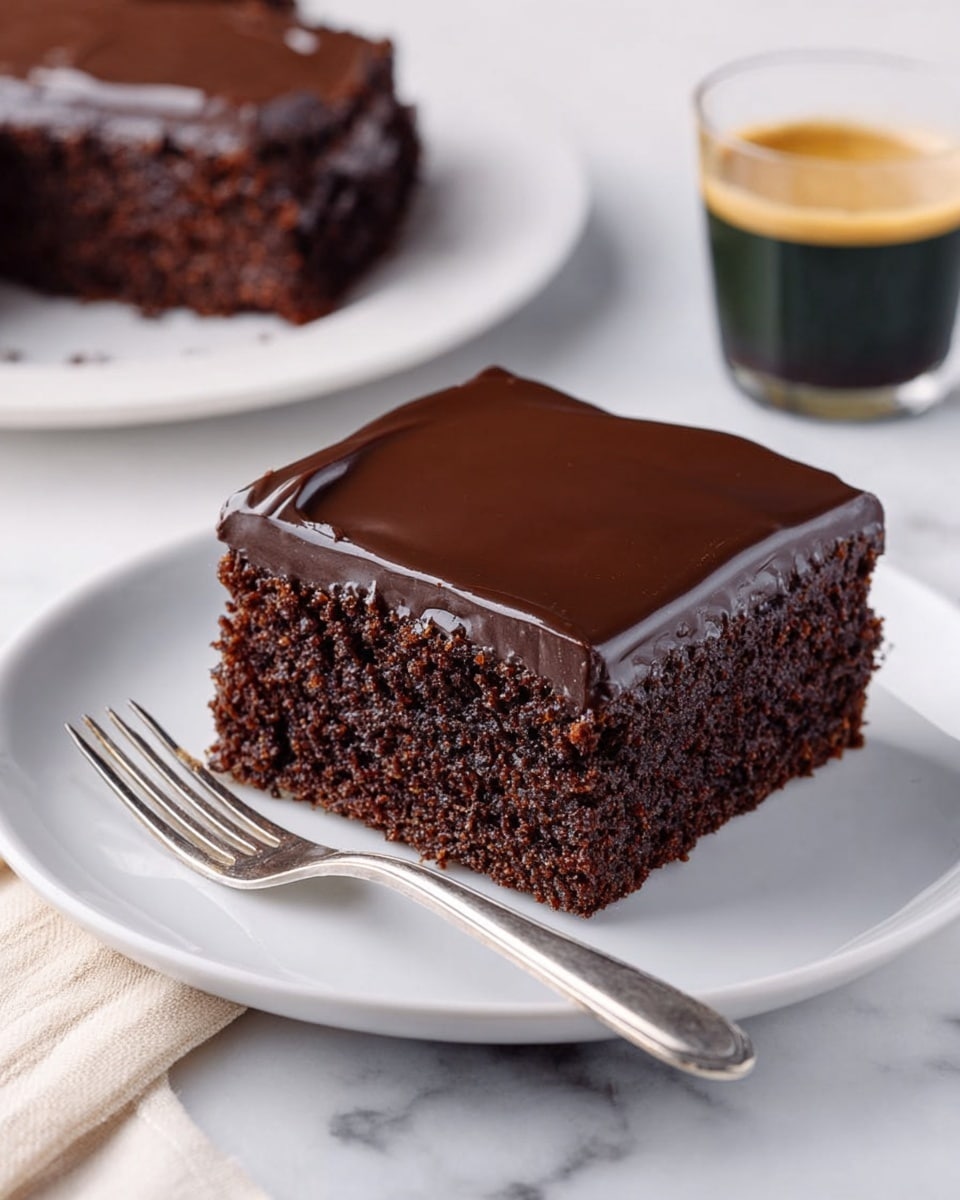 The image shows a square piece of chocolate cake on a white plate placed on a white marbled surface. The cake has two layers: the bottom layer is a thick, dark brown, moist cake with a slightly crumbly texture, while the top layer is a smooth, glossy dark chocolate frosting that covers the whole surface evenly. There is a slight indentation on one side of the frosting, creating a shiny reflection. A silver fork rests near the front edge of the plate and in the background, there is another piece of the cake on a white plate and a small glass of coffee. photo taken with an iphone --ar 4:5 --v 7
